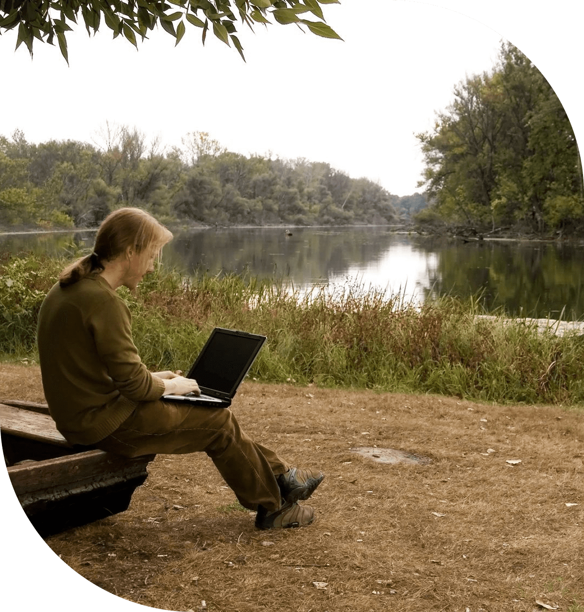Person sitting by a lake using a laptop on a bench.